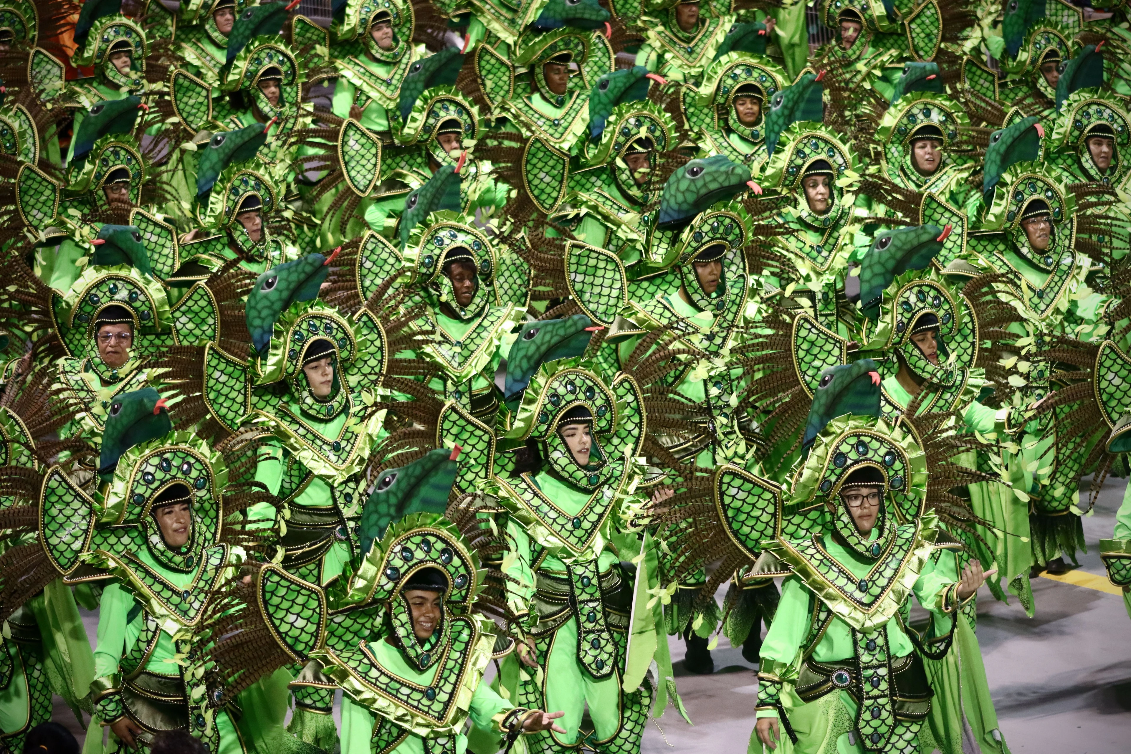 Samba-school participants in patterned green bird costumes move through a São Paulo parade