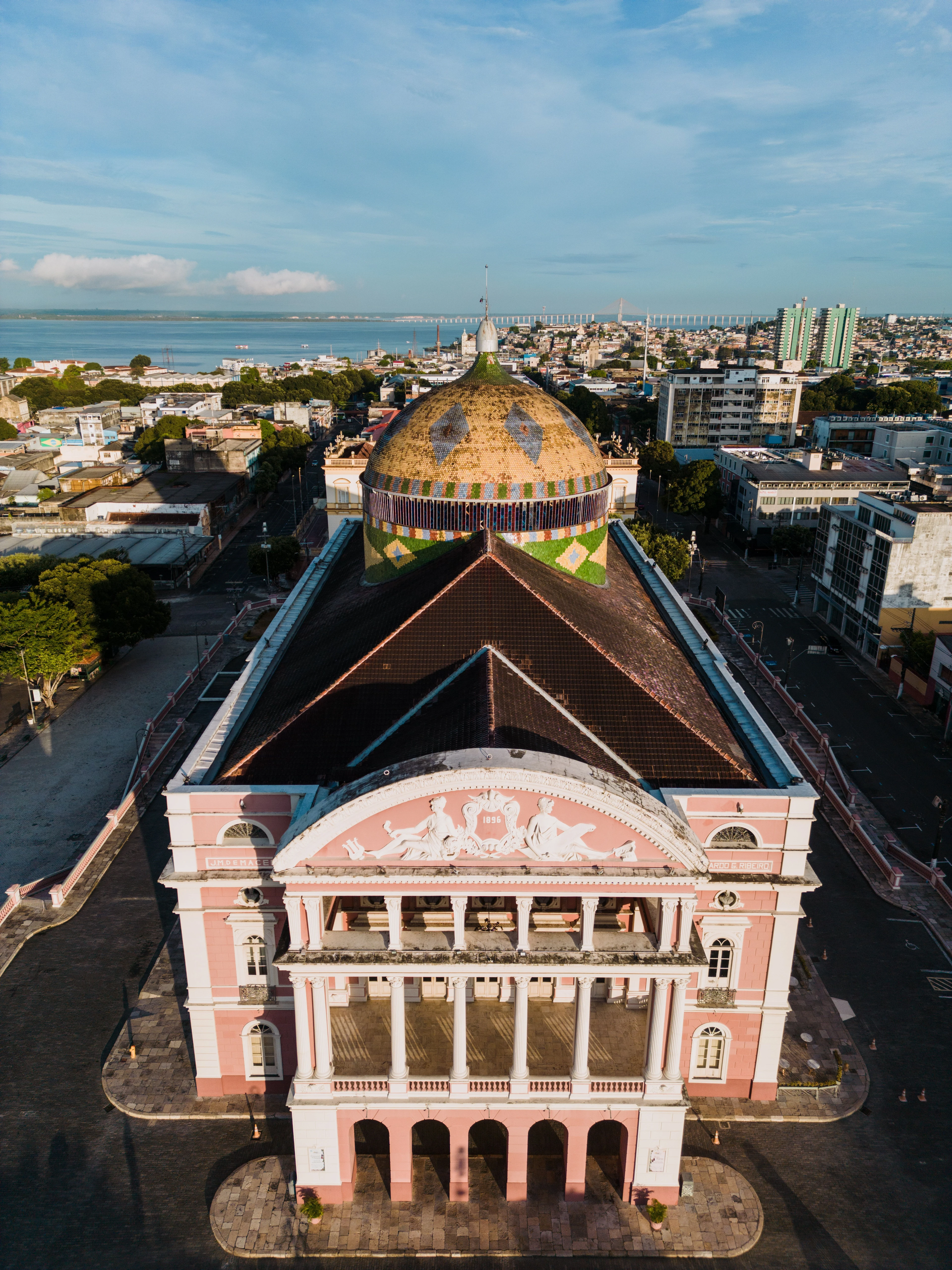 The tiled dome and pink façade of Teatro Amazonas seen from above with Manaus and the river beyond