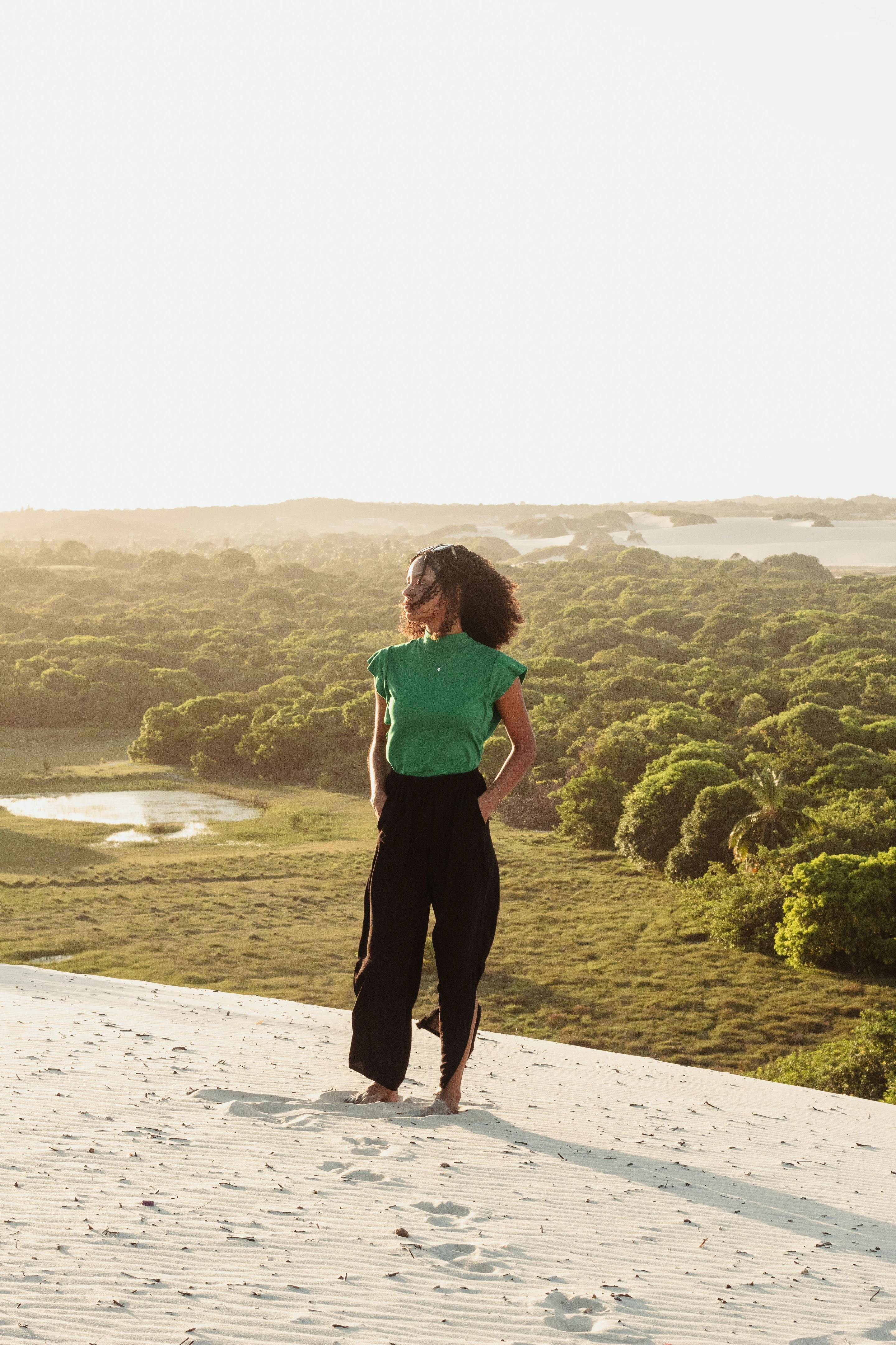 A woman stands on a sunlit sand dune overlooking green vegetation and water in the Natal region