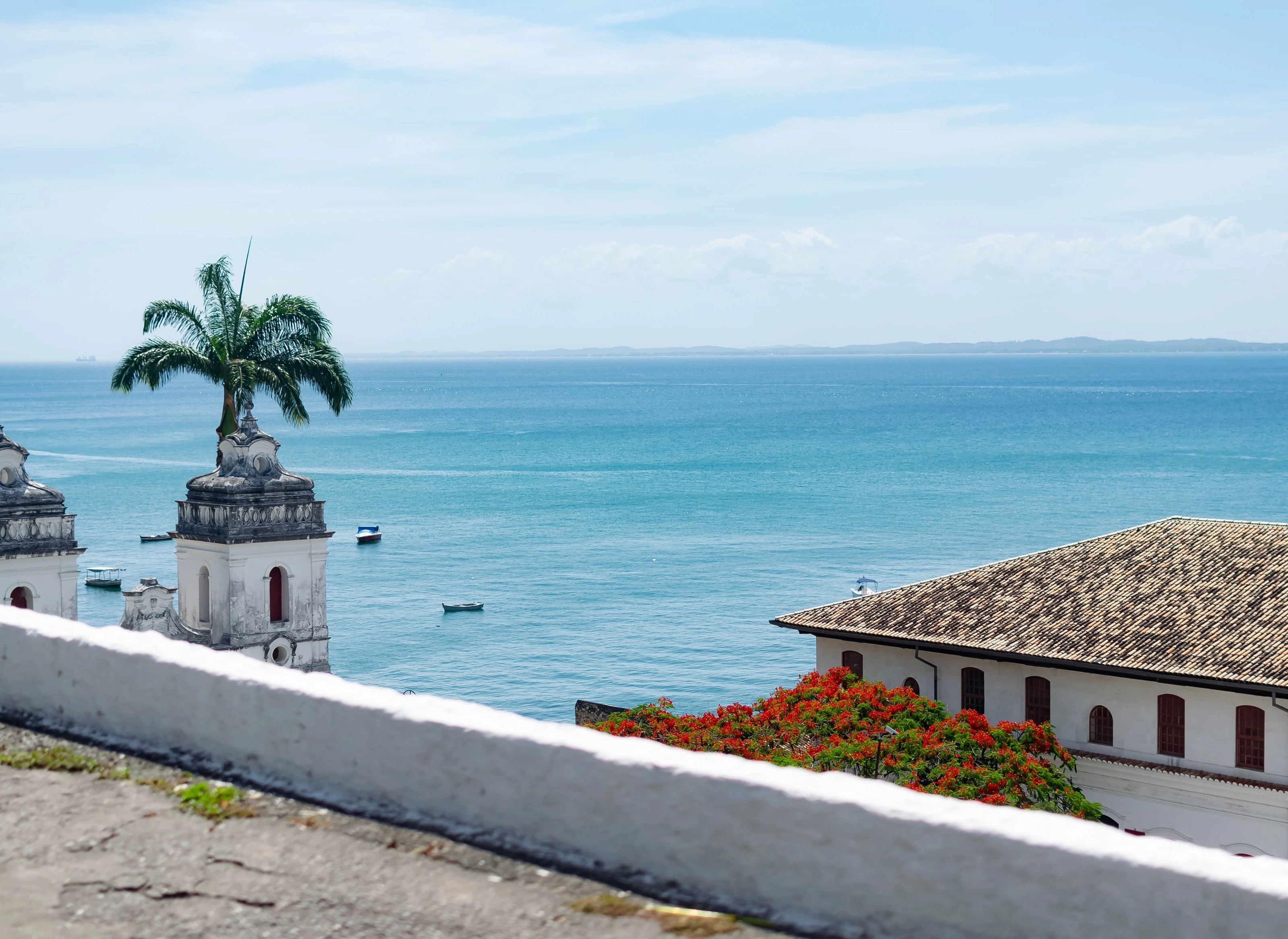 Historic white walls, tiled roofs and red flowers frame a view of Salvador's bay