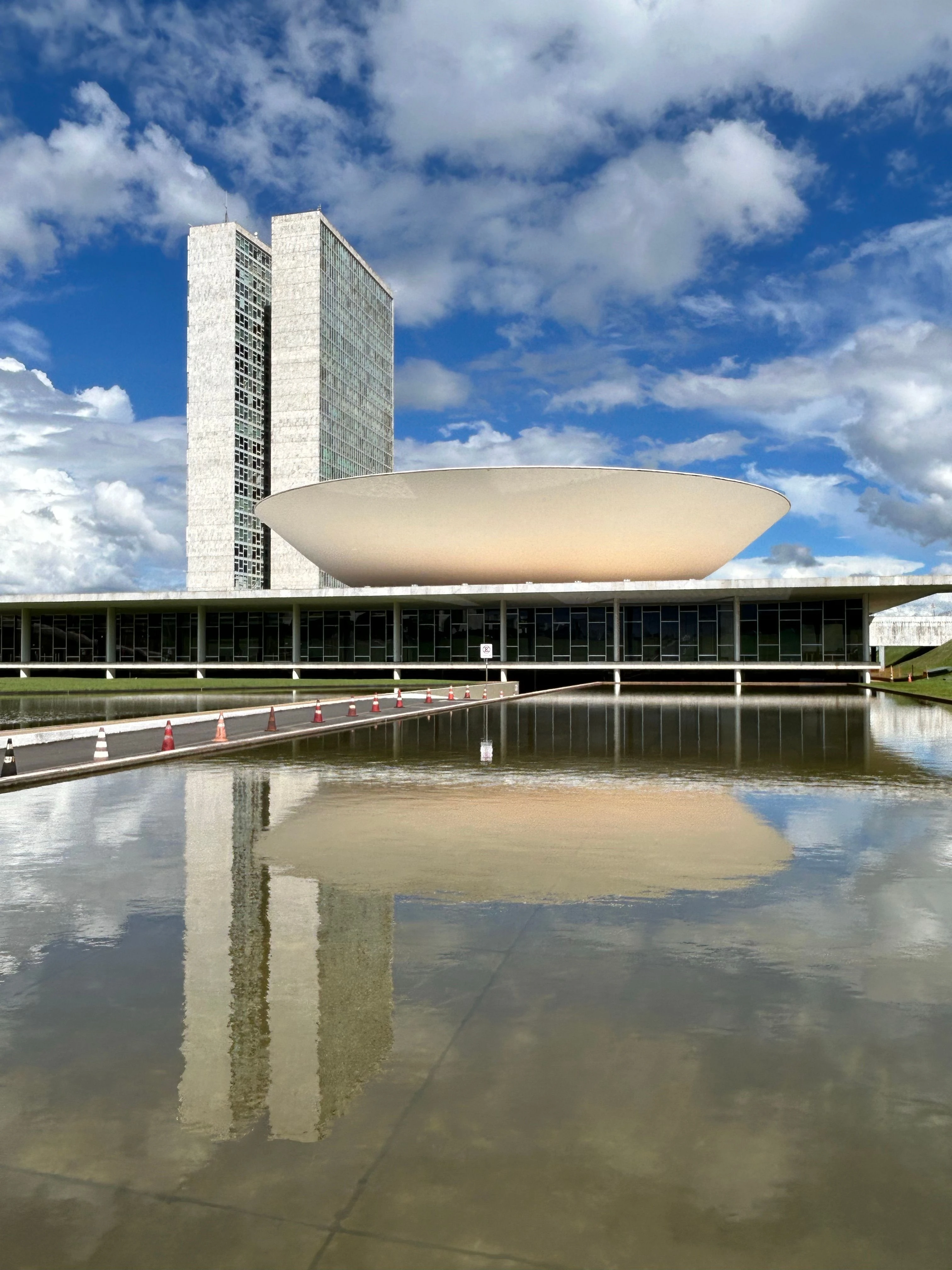 The twin towers and curved chamber of Brazil’s National Congress reflected in water beneath a blue, cloud-filled sky.
