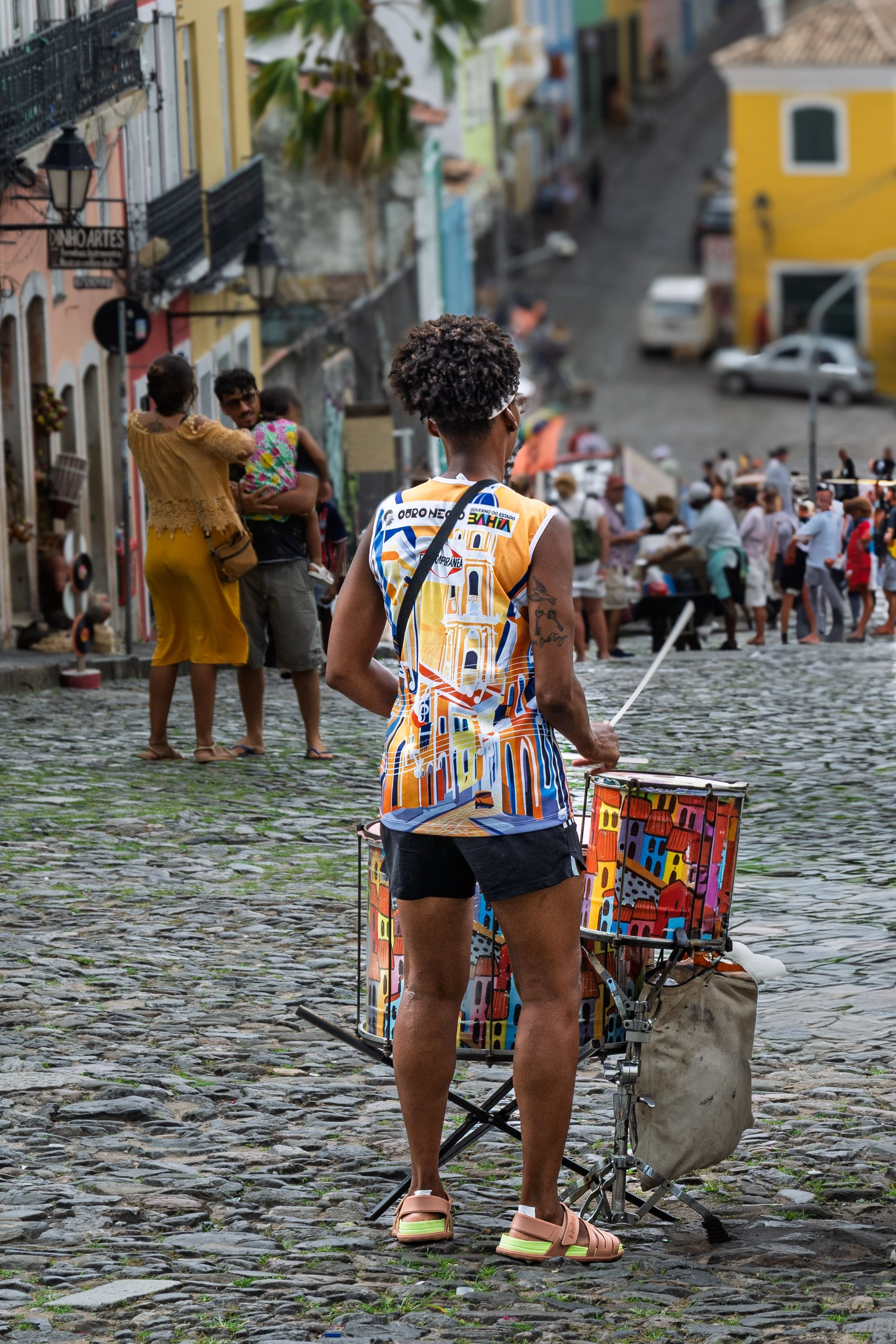 A percussionist seen from behind in a bright shirt plays a drum on a Pelourinho street
