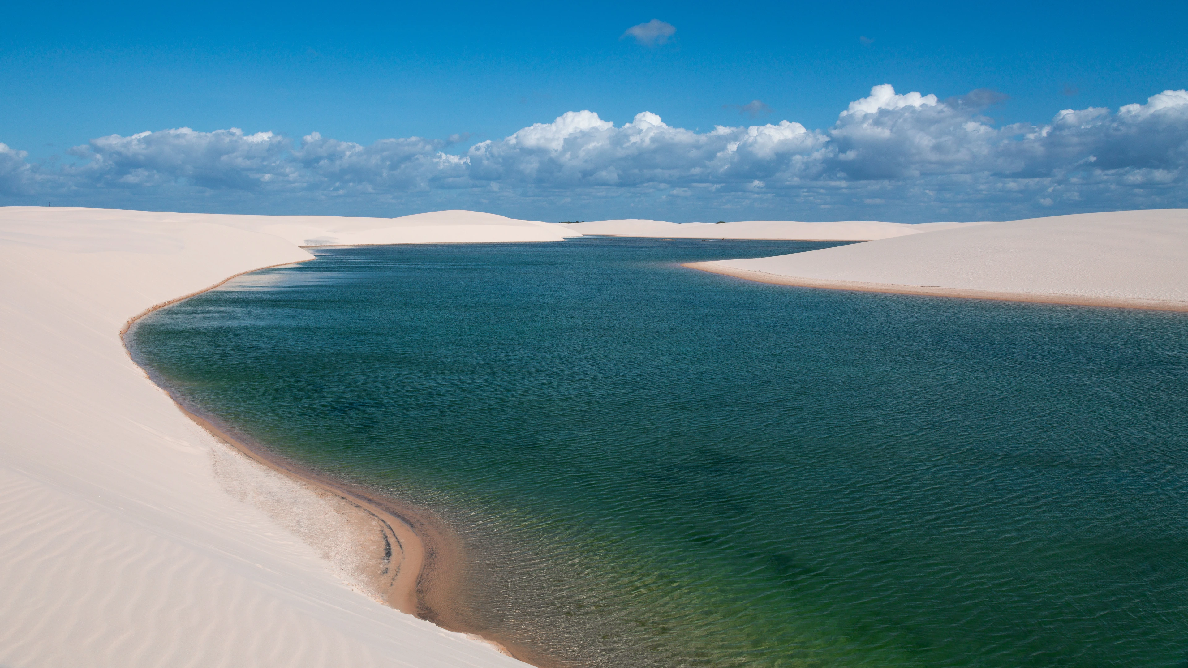 White dunes curving around a deep-blue lagoon beneath bright clouds in Lençóis Maranhenses