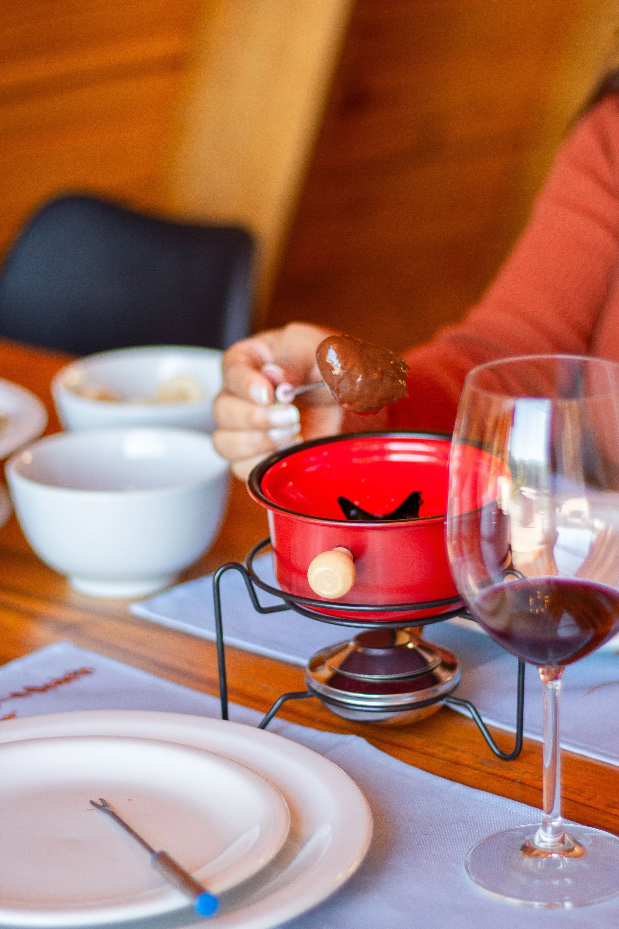 A chocolate-coated piece of fruit is lifted above a red fondue pot beside wine in Serra Negra, Brazil.