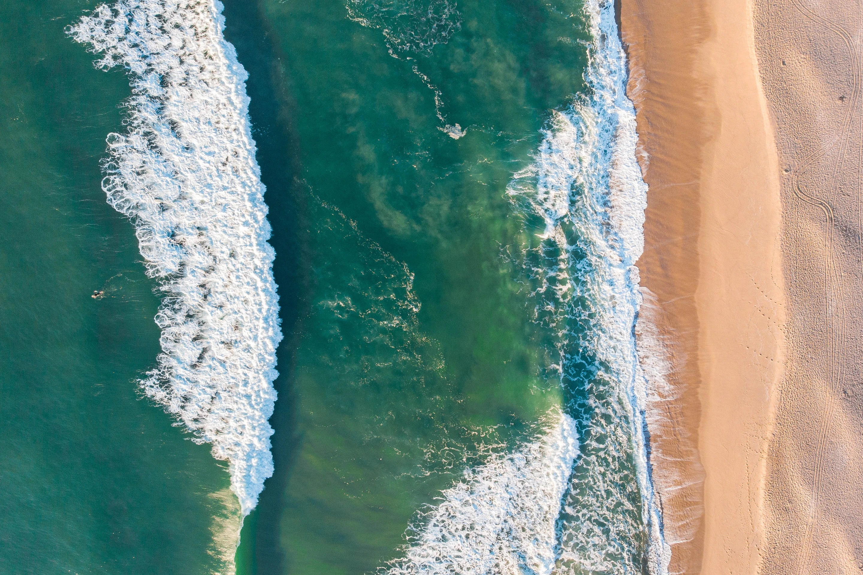 White surf traces sweeping lines across emerald Atlantic water and warm sand in an overhead view of Florianópolis.