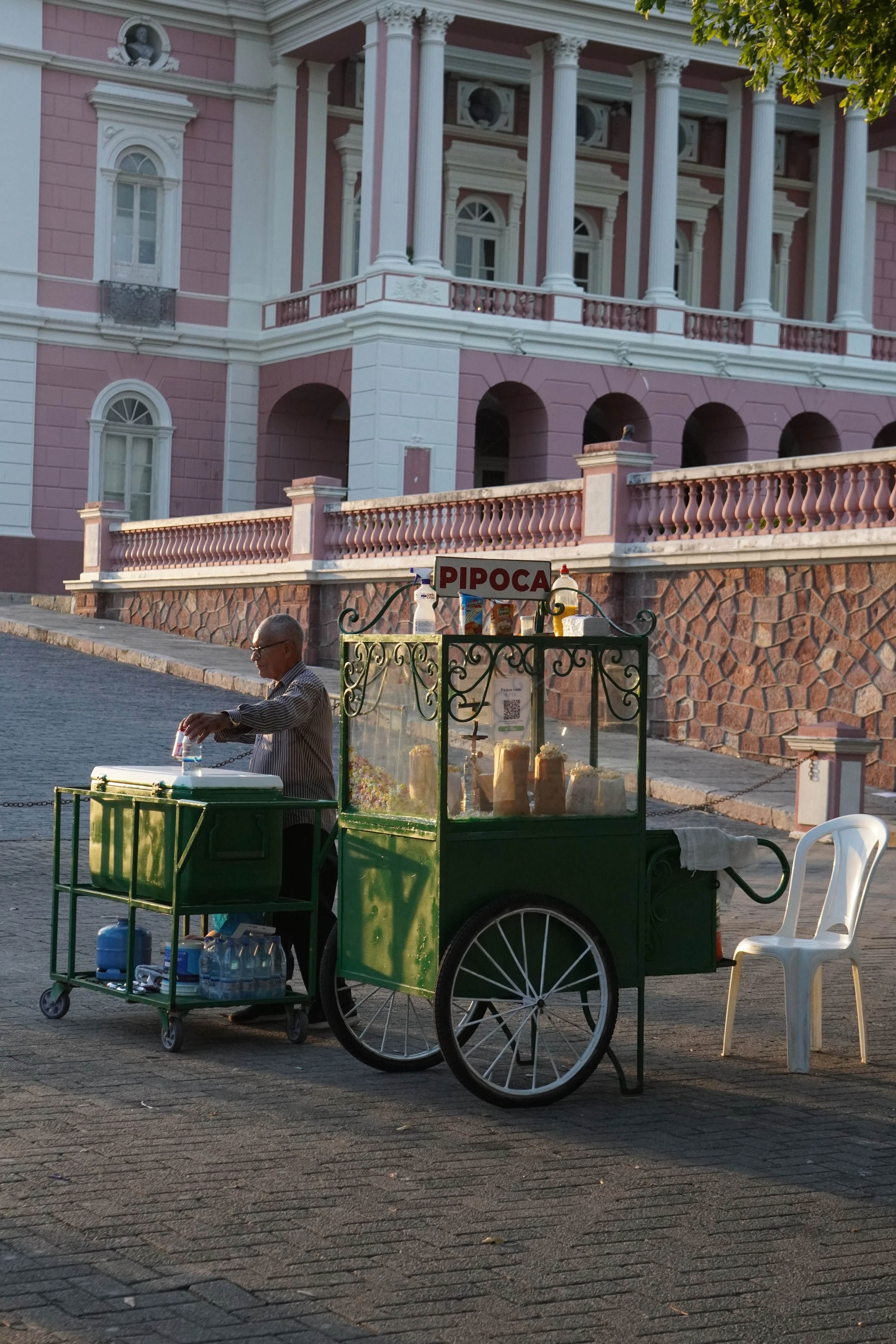 A popcorn vendor's colorful cart stands before the pink façade and dome of Teatro Amazonas