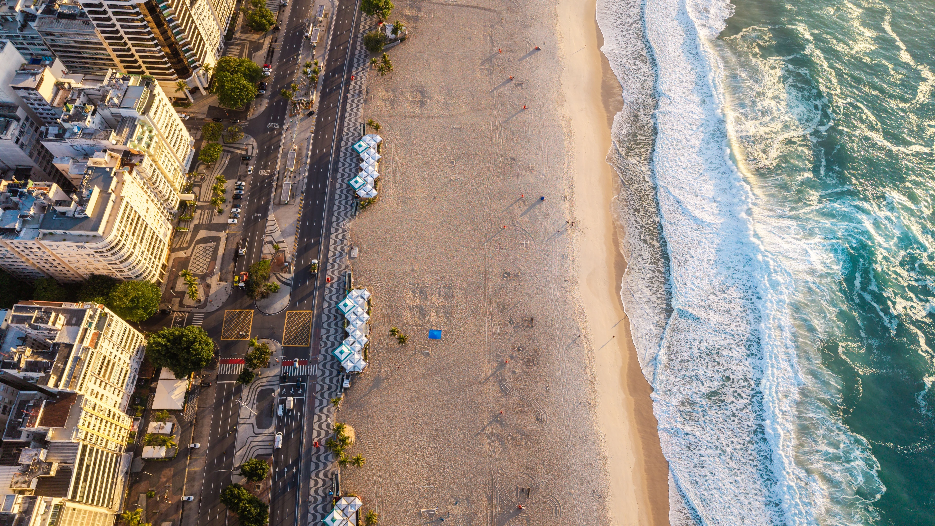 An overhead view of Copacabana’s patterned promenade, sunlit buildings and breaking Atlantic waves.