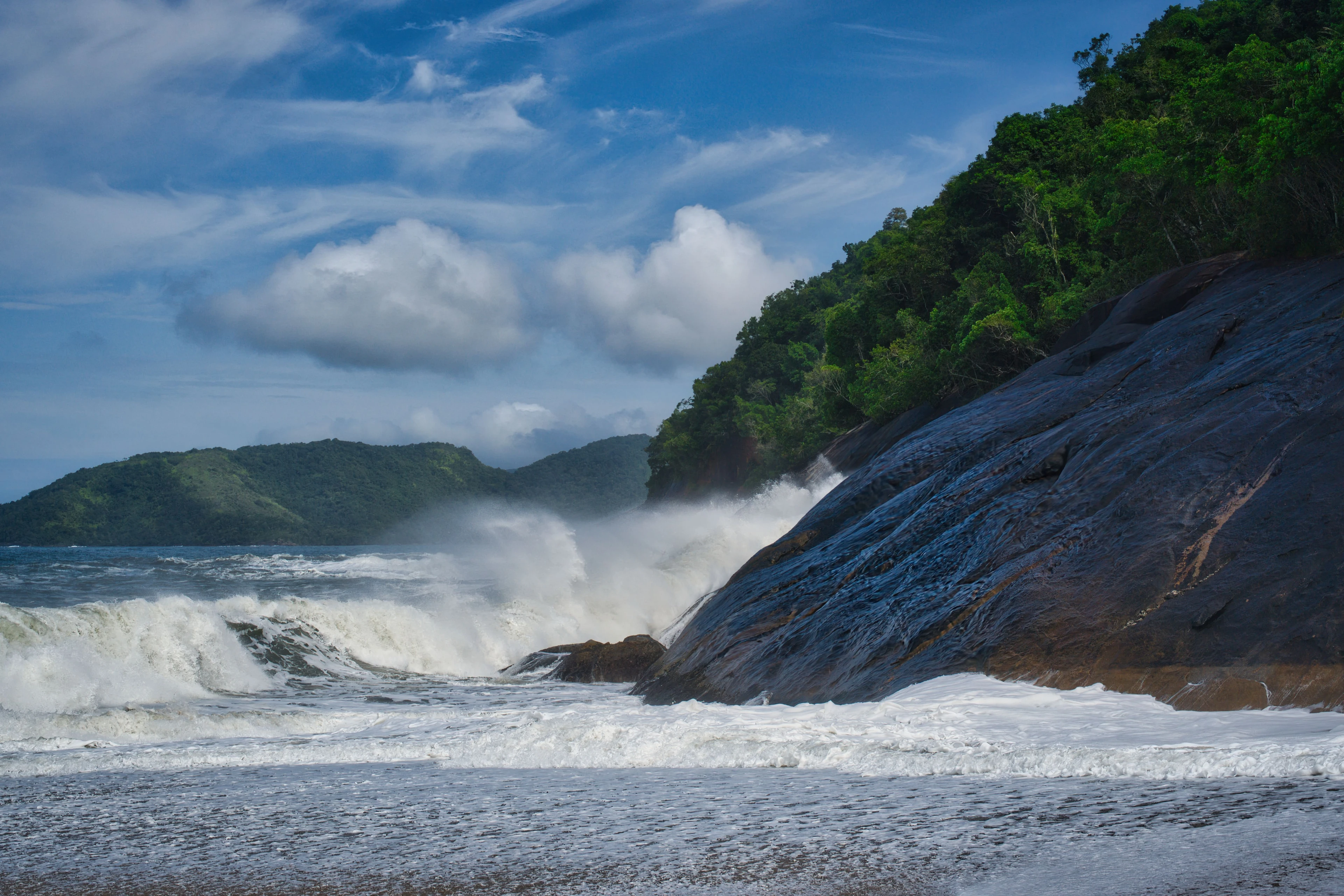 Powerful white waves breaking against a smooth forest-backed rock face