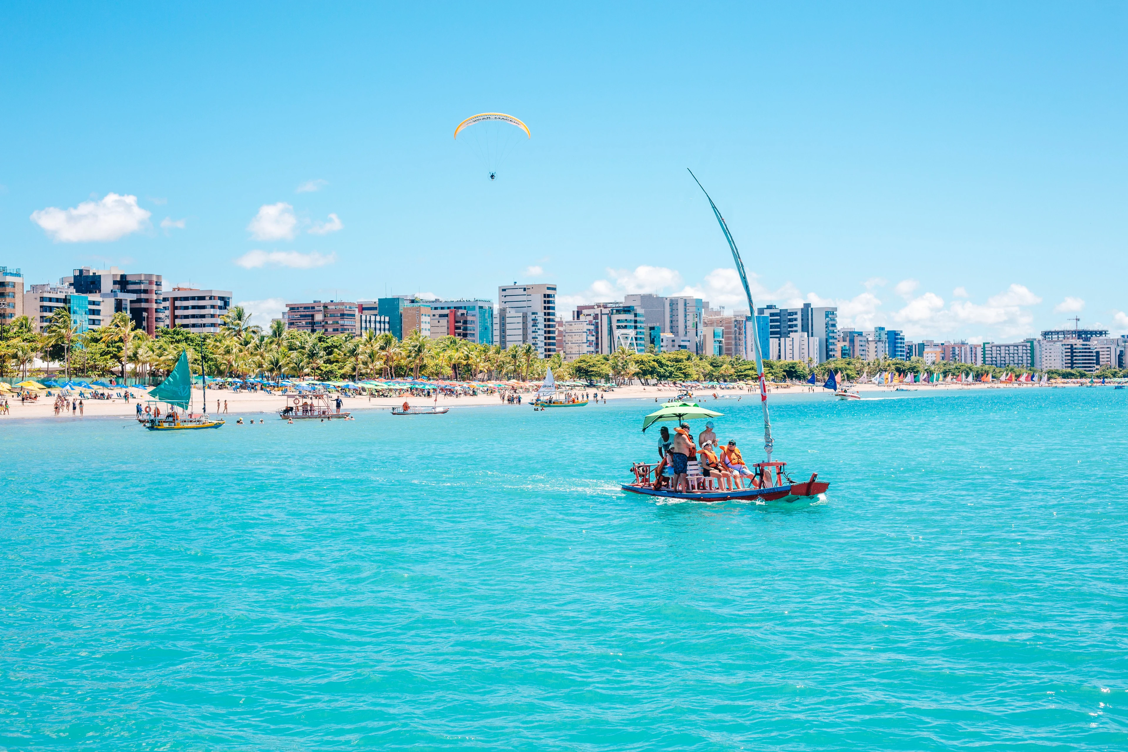 A jangada carrying people on vivid blue water with Maceió’s active Pajuçara waterfront behind