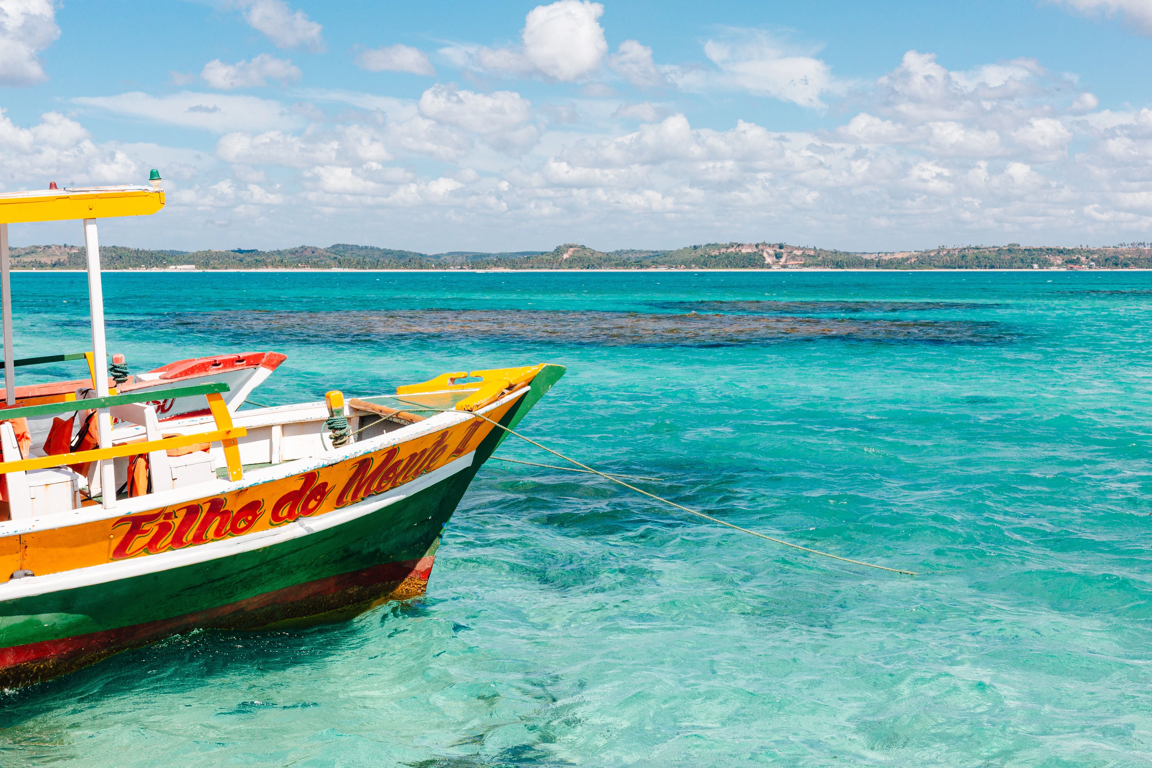 A brightly painted yellow, red and green boat floats over transparent reef water at Maragogi
