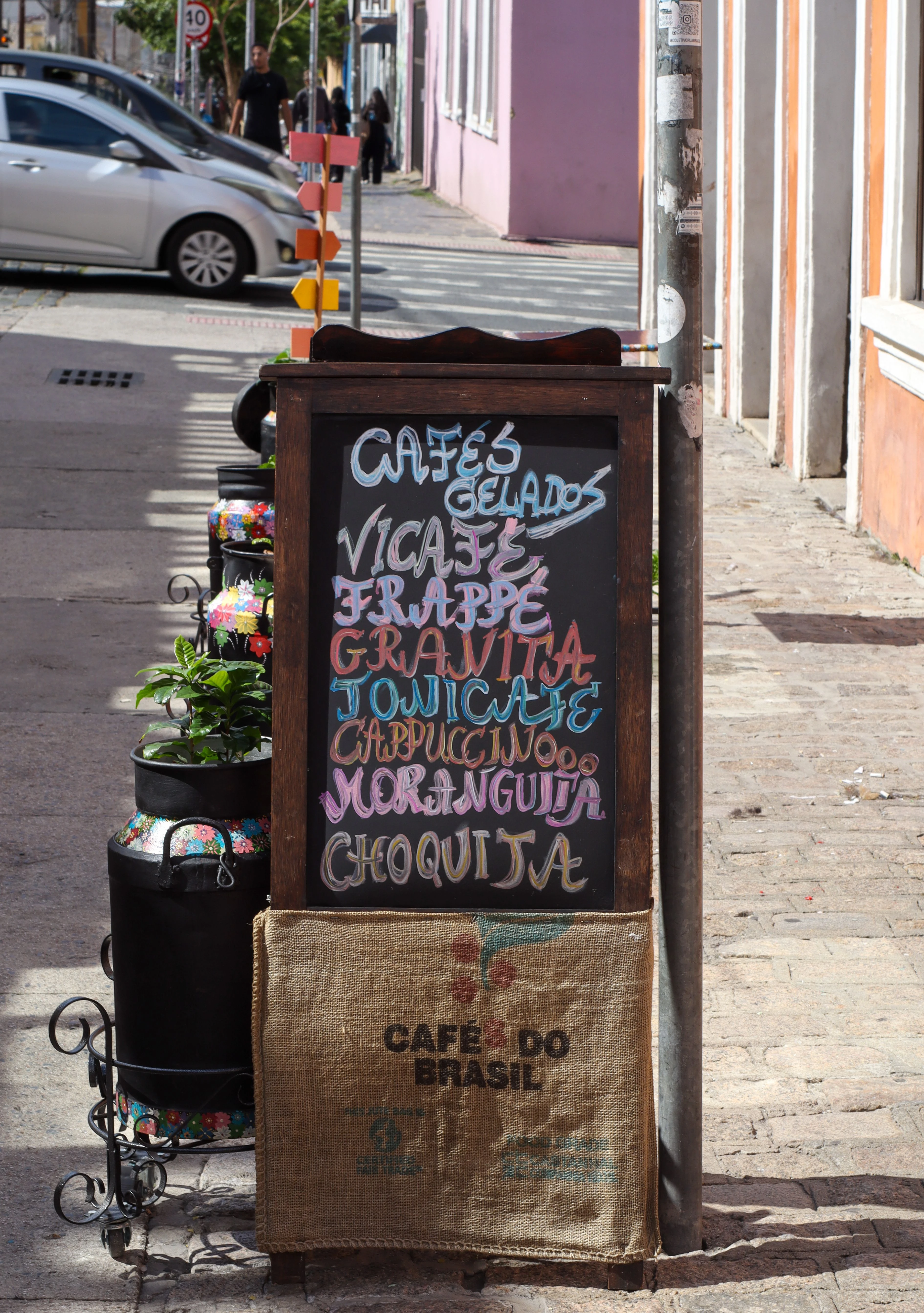 A café's colorful chalkboard lists drinks in Portuguese beside a Brazilian sidewalk