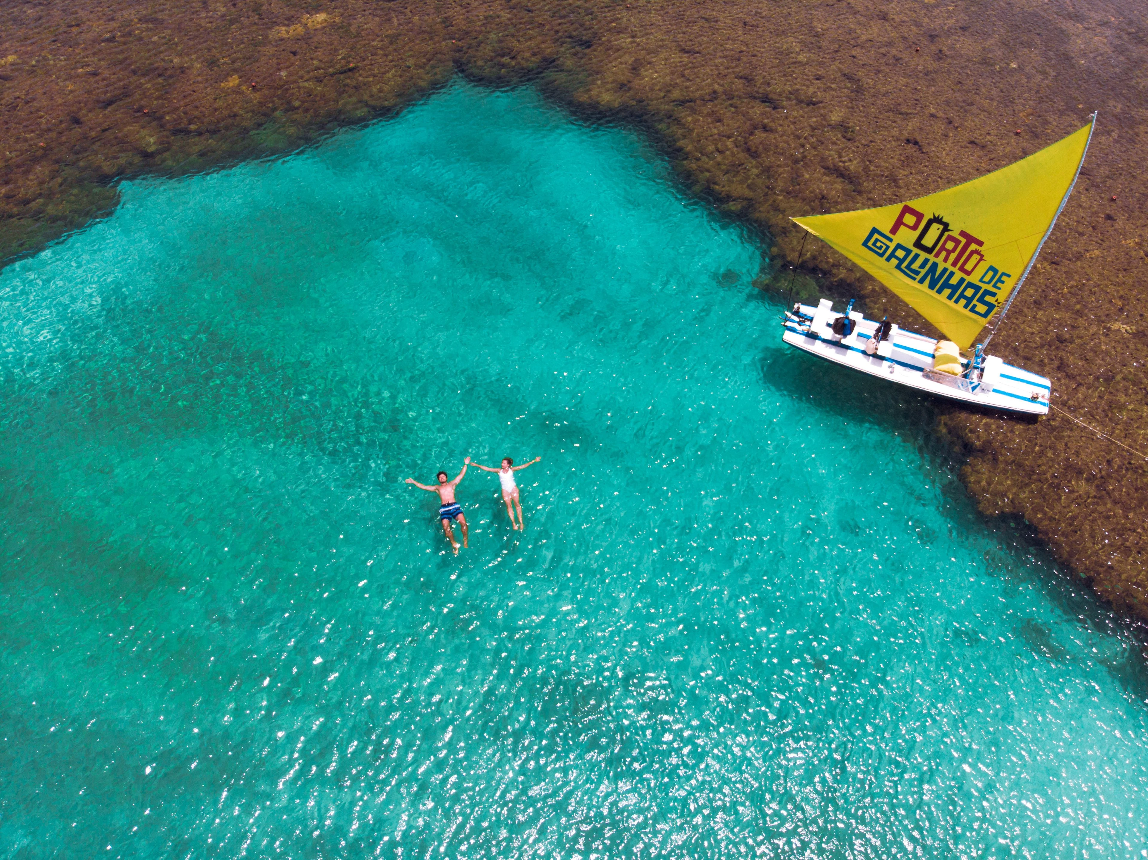 A yellow-sailed jangada and swimmers seen overhead in clear turquoise water beside a reef at Porto de Galinhas