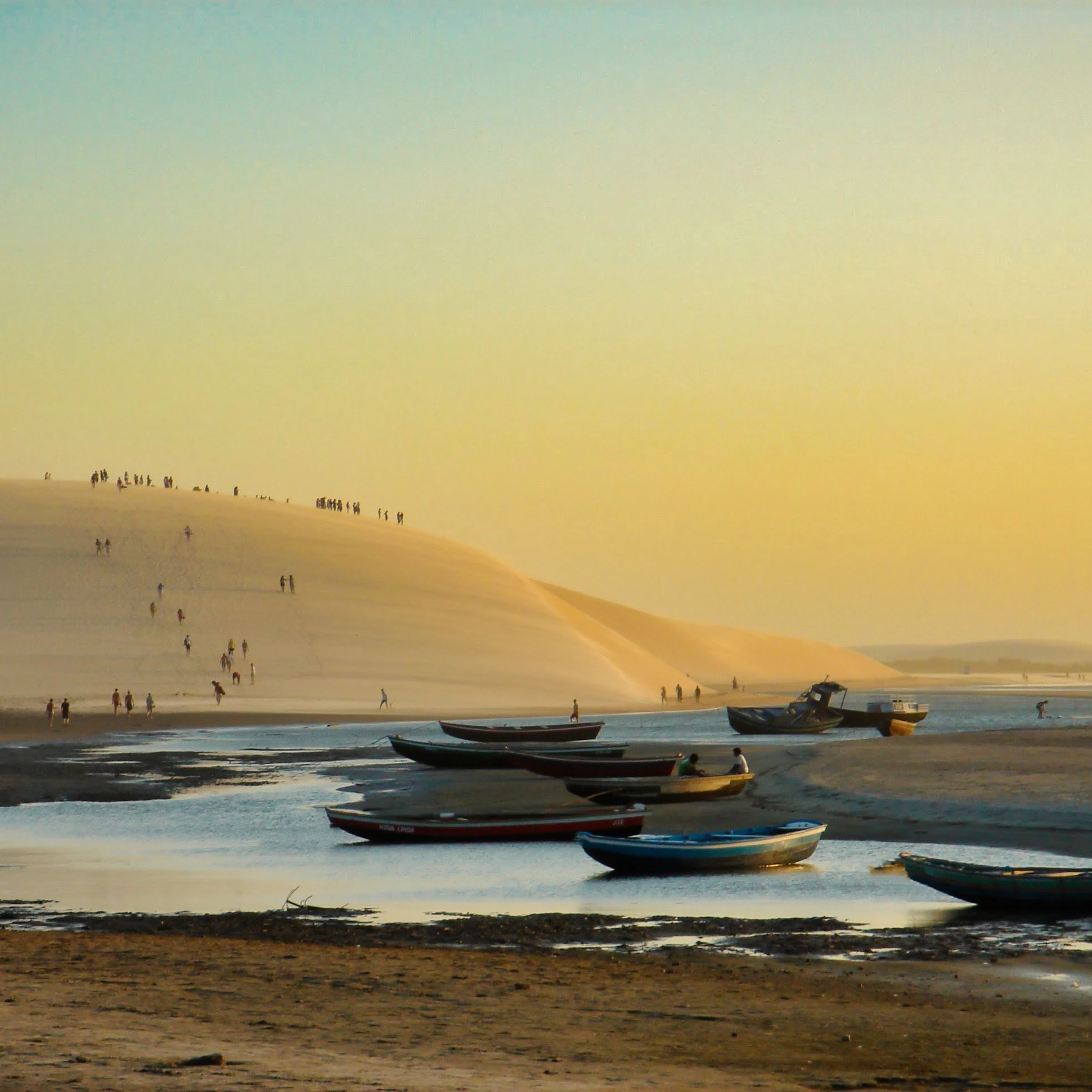 Fishing boats rest near the shore below Jericoacoara’s dunes in golden evening light.