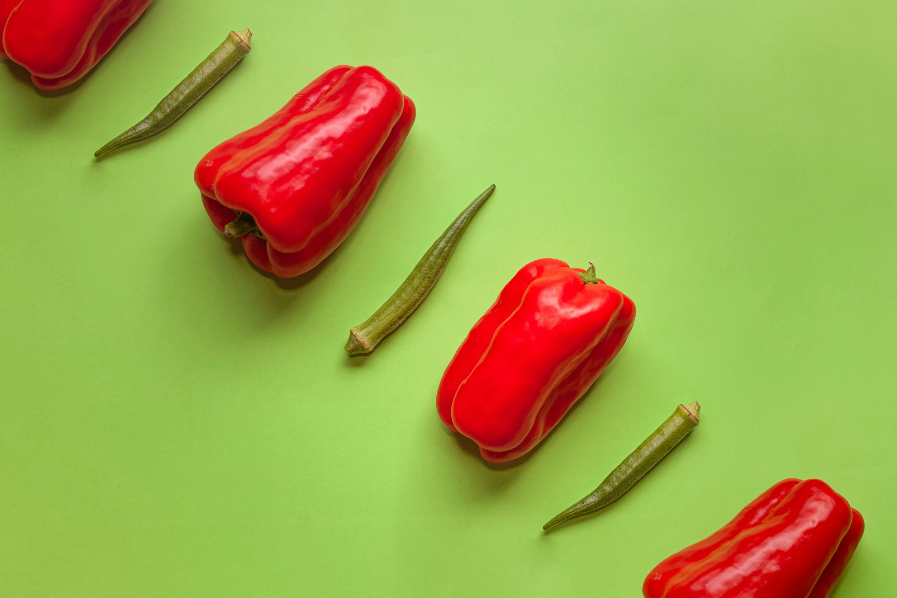 Red bell peppers and green okra arranged diagonally across a green surface